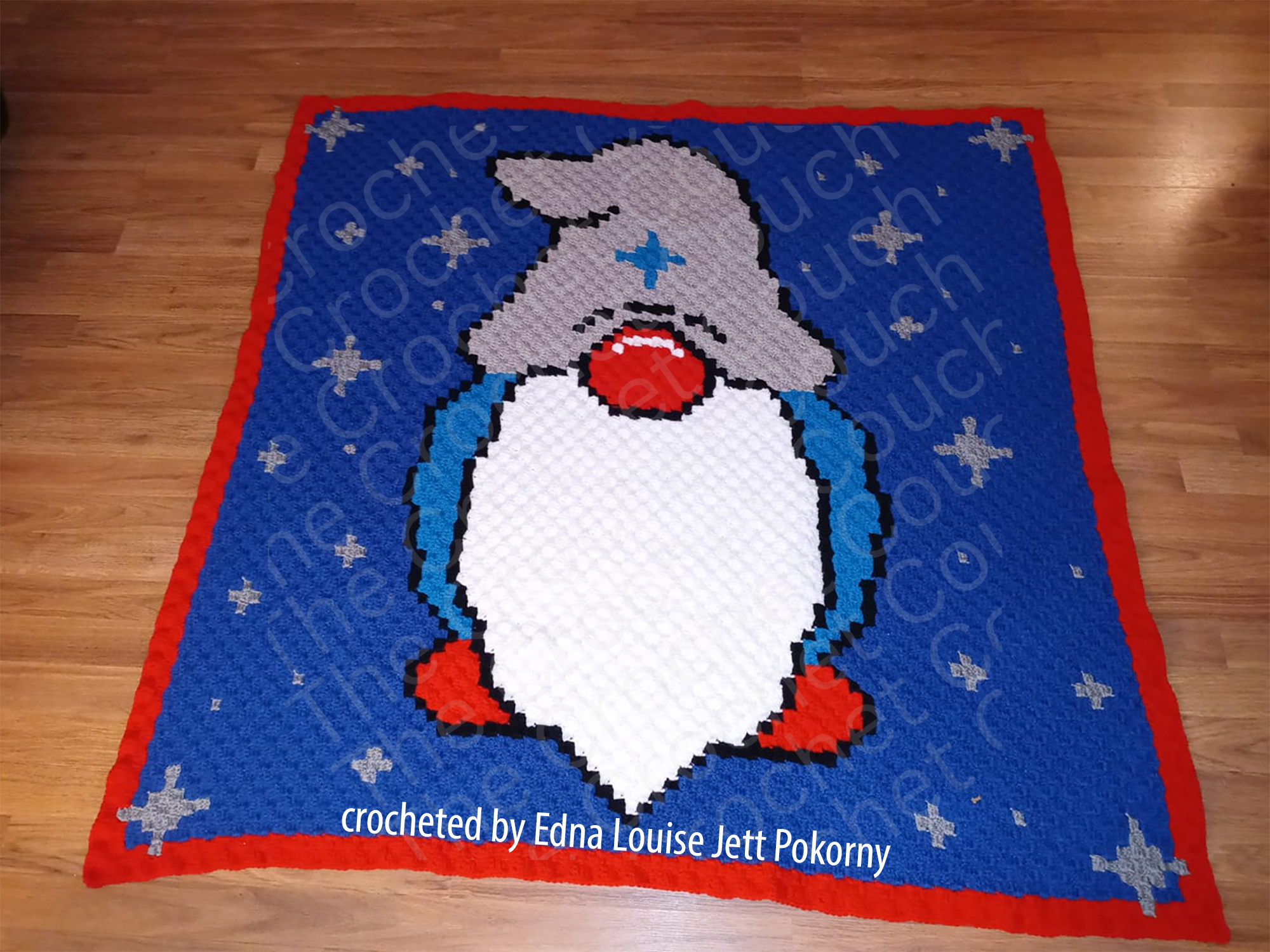 A handmade crochet afghan featuring a whimsical gnome with a gray hat and blue outfit, set against a blue background with stars and a red border, laid out on a wooden floor.