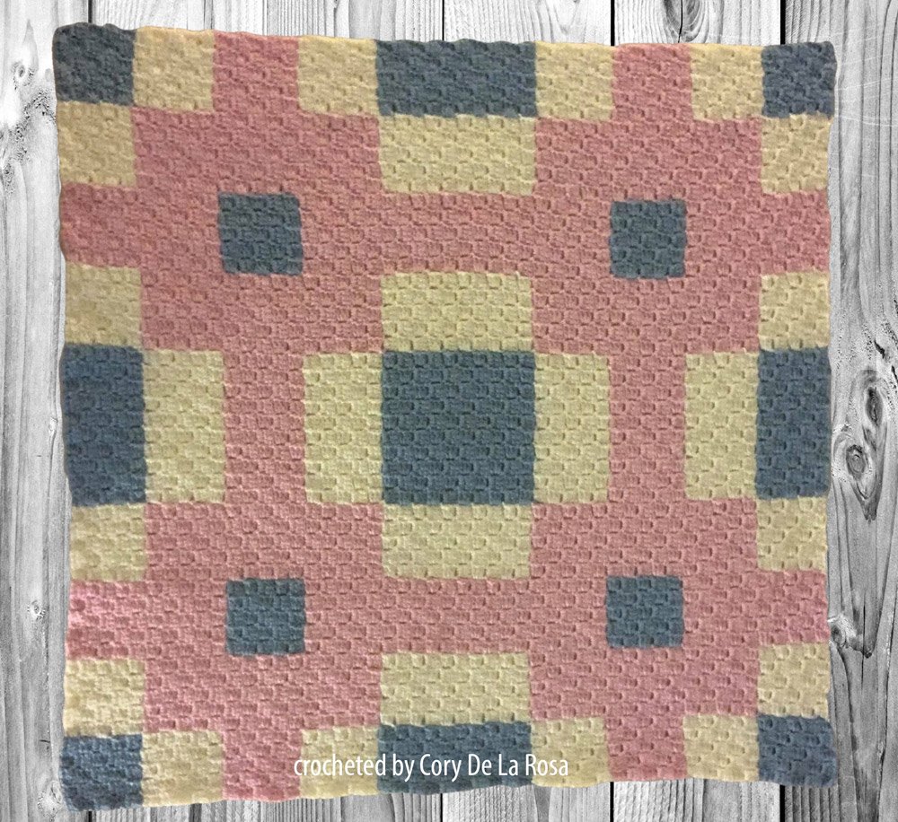 Crocheted afghan in a quilt-inspired design with soft pink, yellow, and blue blocks arranged in a cozy geometric pattern.