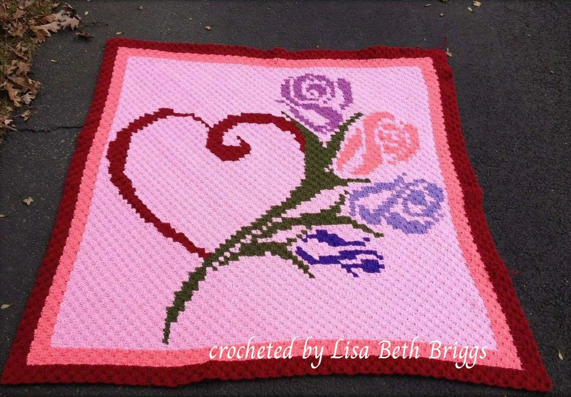 A crochet afghan in C2C style with a large sweeping heart in red and roses in purple, lavender, and peach on a pink background with a red border.