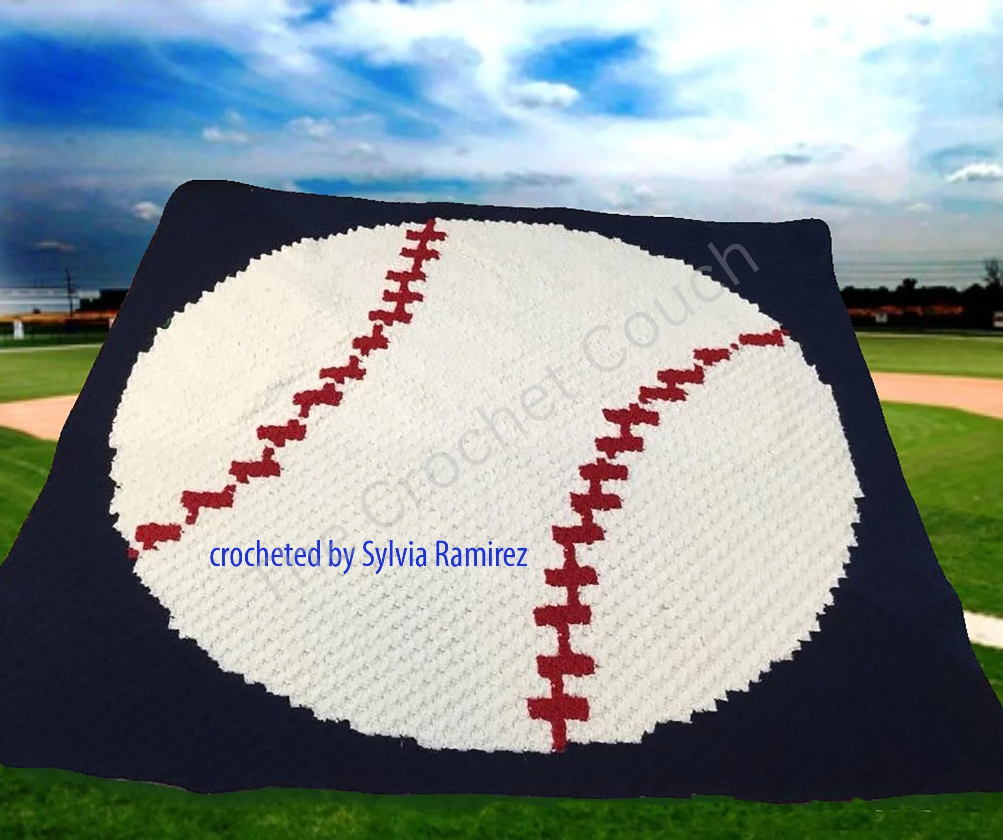 A crocheted afghan with a large white baseball design featuring red stitching on a dark background, made by Sylvia Ramirez. Displayed against a baseball field background.
