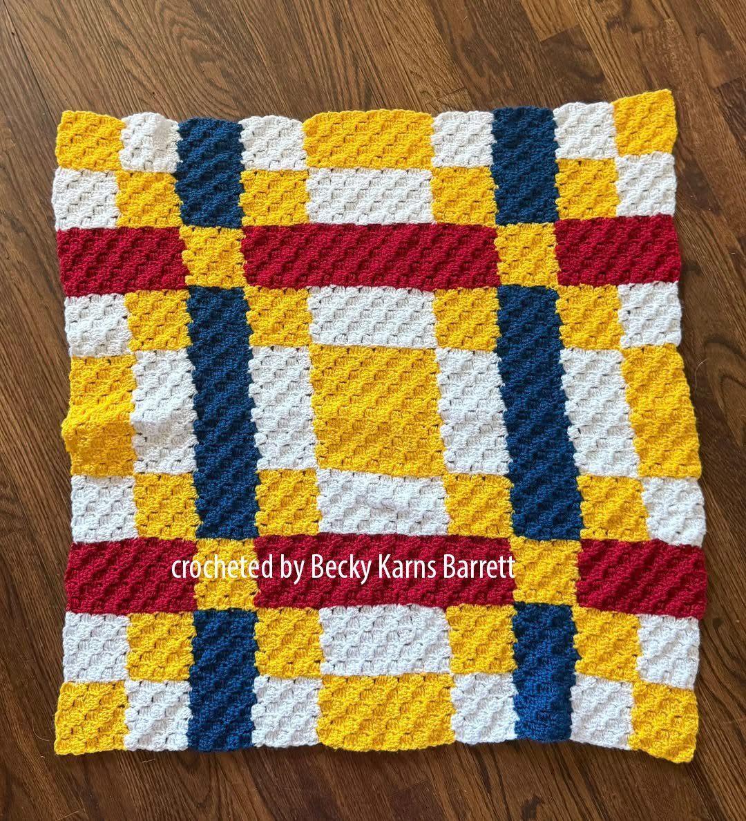 Corner-to-corner crochet afghan laid on a wood floor, featuring a quilt-inspired plaid pattern in blocks of golden yellow, white, navy, and red.