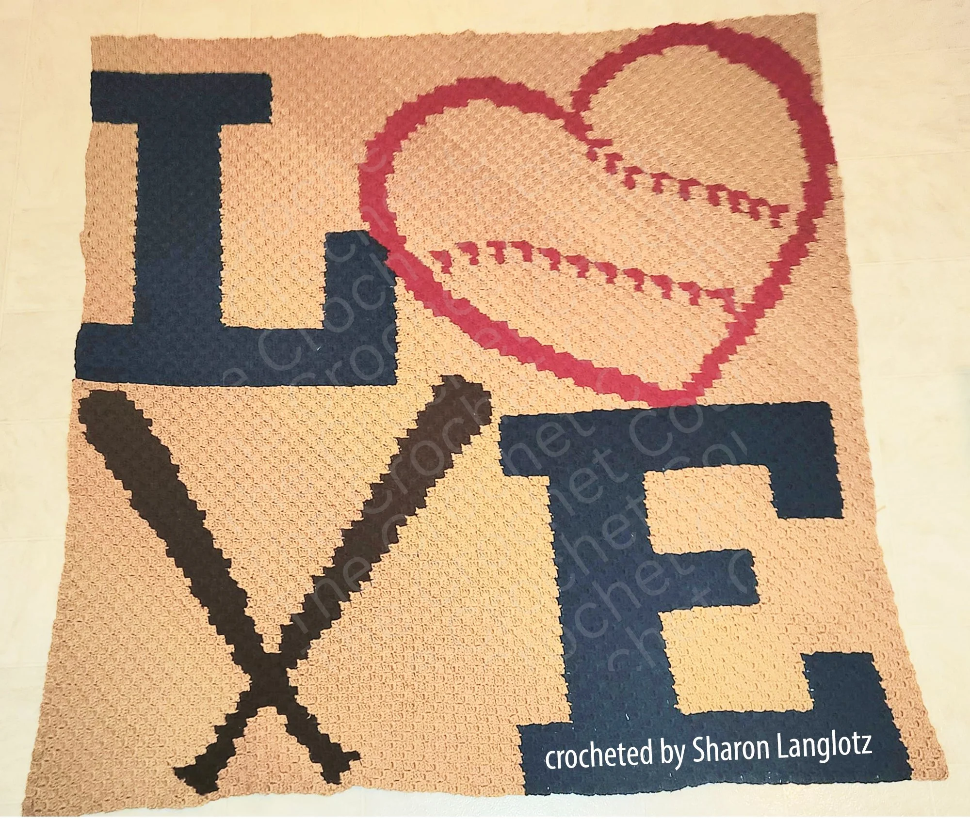 A handmade C2C crochet afghan featuring the word 'LOVE' with a heart-shaped baseball and crossed bats, designed in shades of blue, red, and beige, laid flat on the floor.