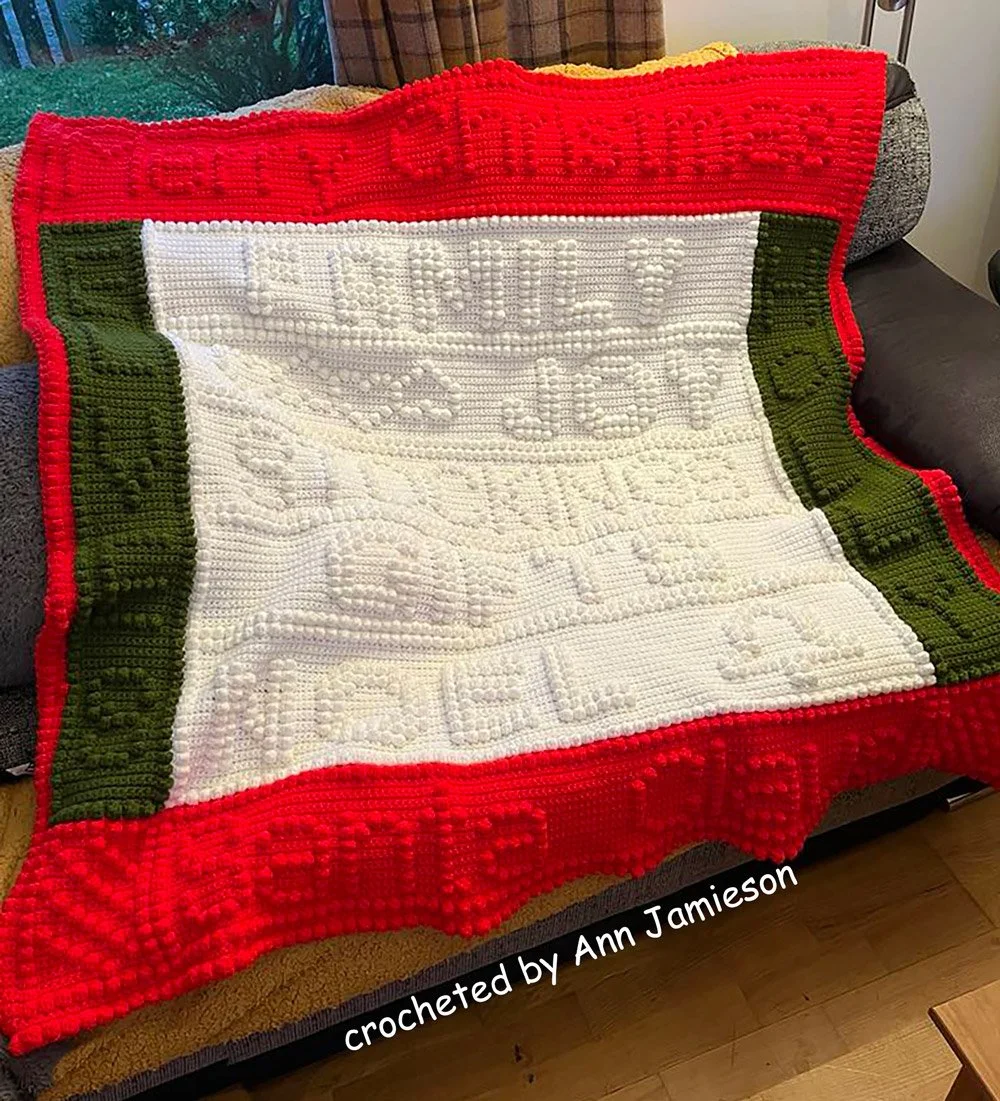 A cozy Christmas afghan featuring classic holiday motifs in red, green, and white, adding festive charm to a living room. Crocheted by Ann Jamieson.