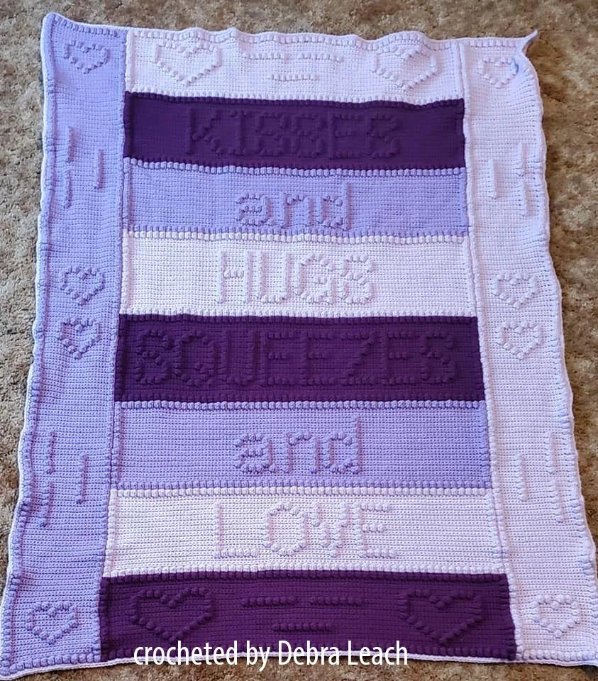 A crochet afghan in shades of purple, featuring embossed words that read "Kisses and Hugs Squeezes and Love," with heart details along the border. A cozy and sentimental handmade blanket.