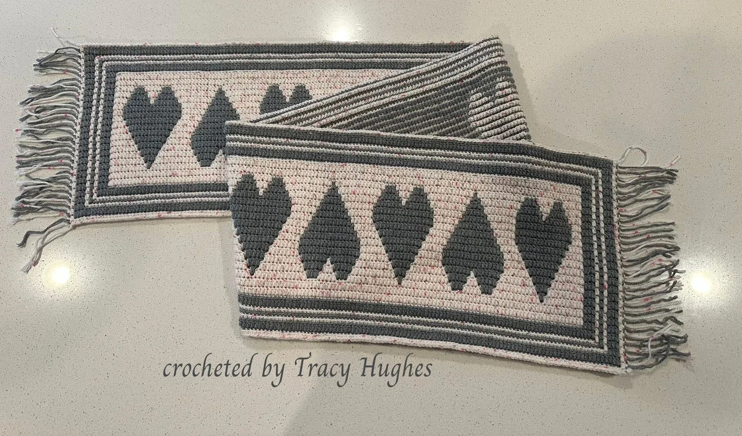 Crocheted table runner with five large gray hearts centered in a soft speckled pink background, bordered by bold gray and pink stripes with fringe on both ends. Displayed on a white countertop.