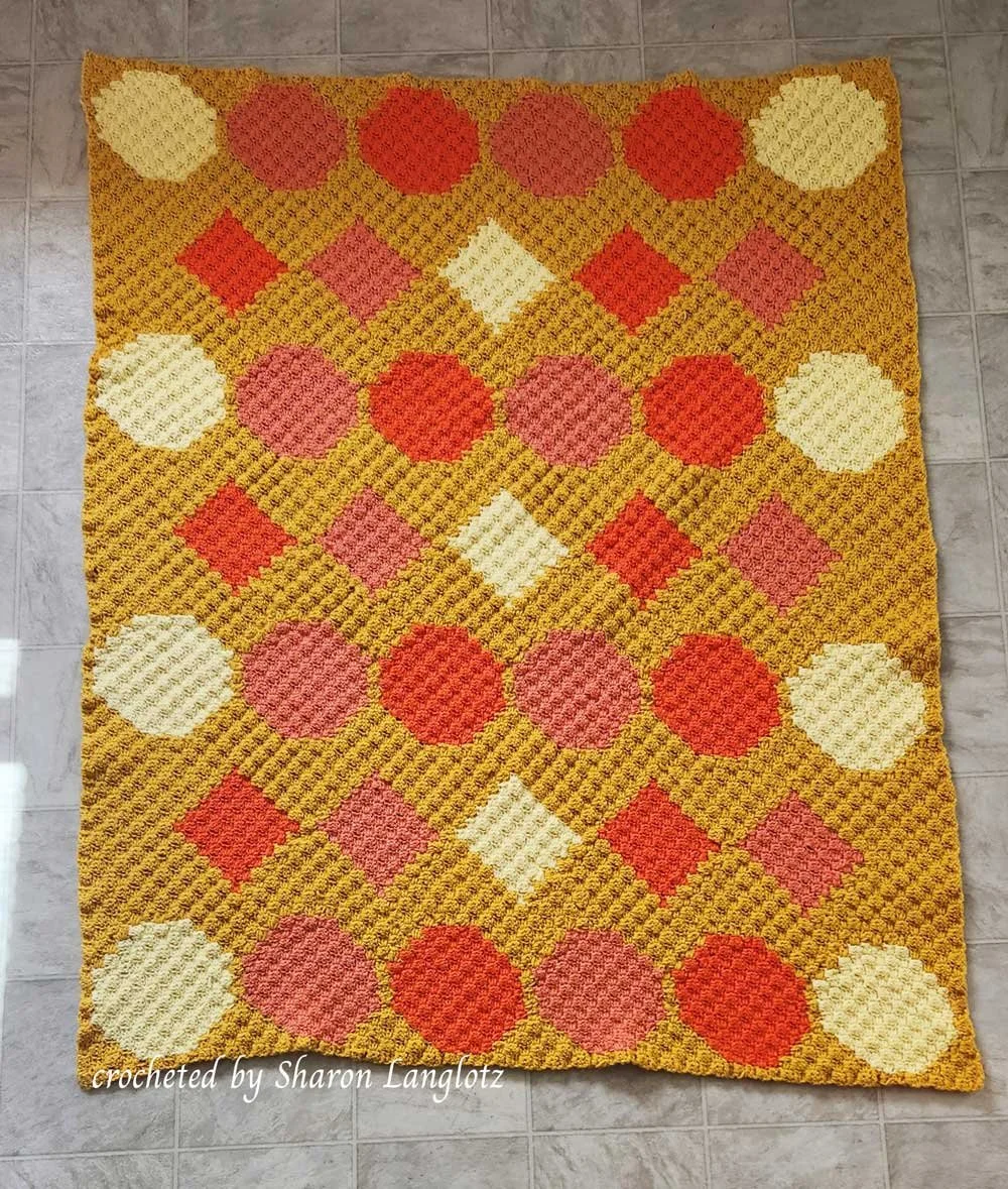 Crocheted afghan featuring bright geometric shapes in red, pink, cream, and yellow on a mustard yellow background