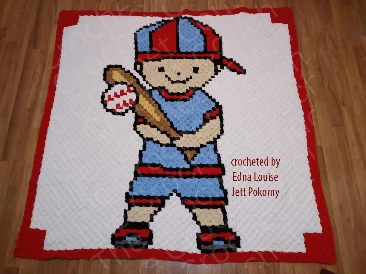 Handmade crochet afghan featuring a baseball boy in red, white, and blue, holding a bat and ball, displayed on a wooden floor.