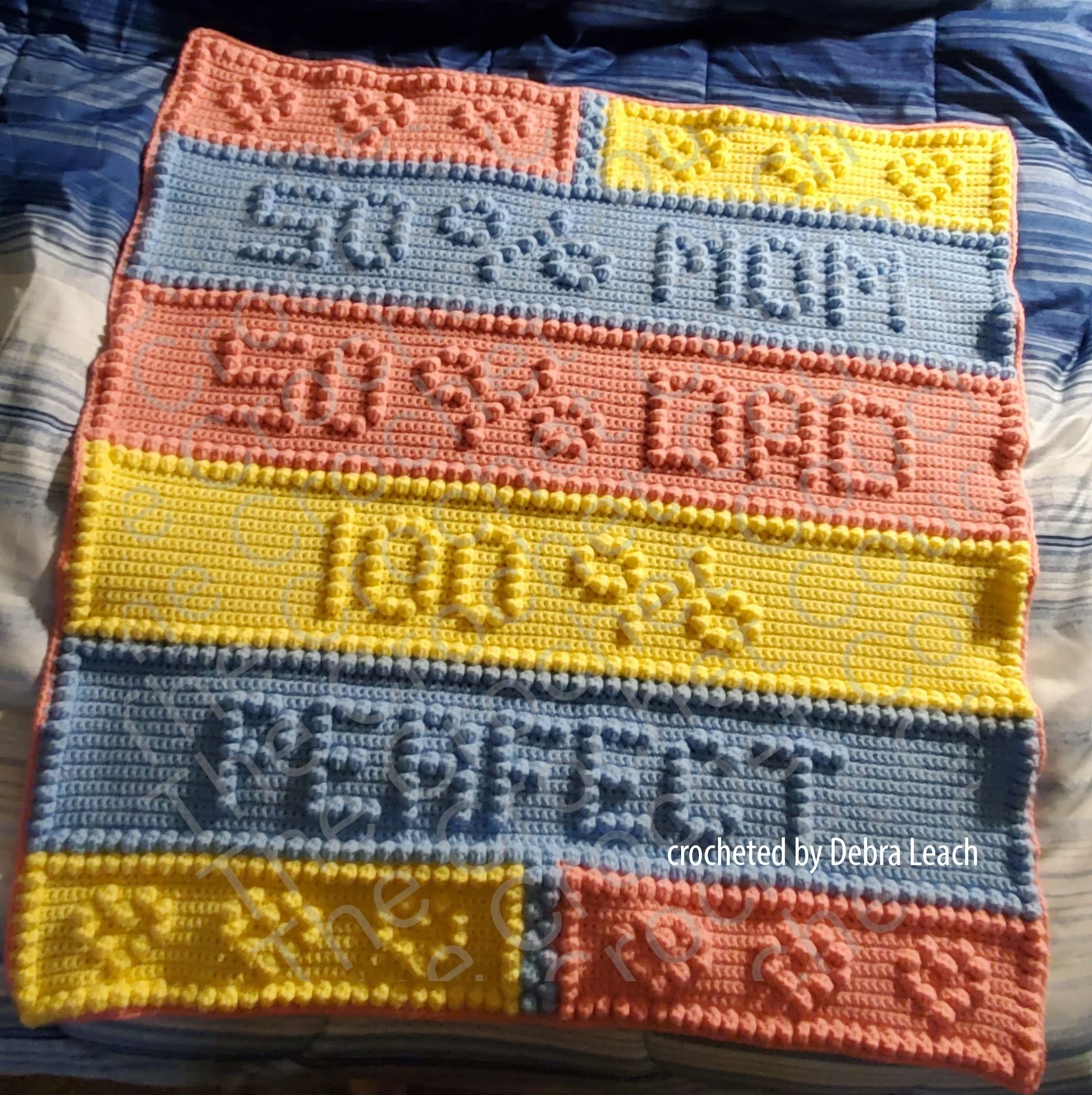 Handmade baby afghan with the phrase "50% Mom, 50% Dad, 100% Perfect" in Bobble stitch, crafted in pastel pink, blue, and yellow stripes, displayed on a beige carpet.