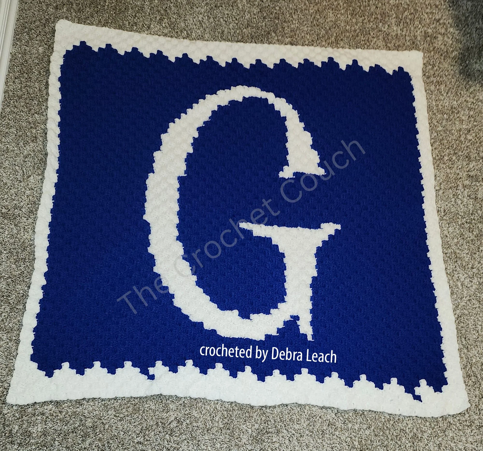 A crochet afghan featuring a large, bold white letter G centered on a deep blue background with a white border.