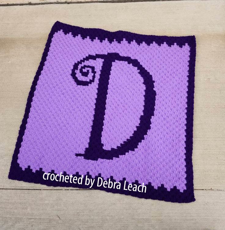 Purple crochet afghan featuring a large, whimsical dark purple letter "D" with a spiral detail at the top left, framed by a matching dark purple border.