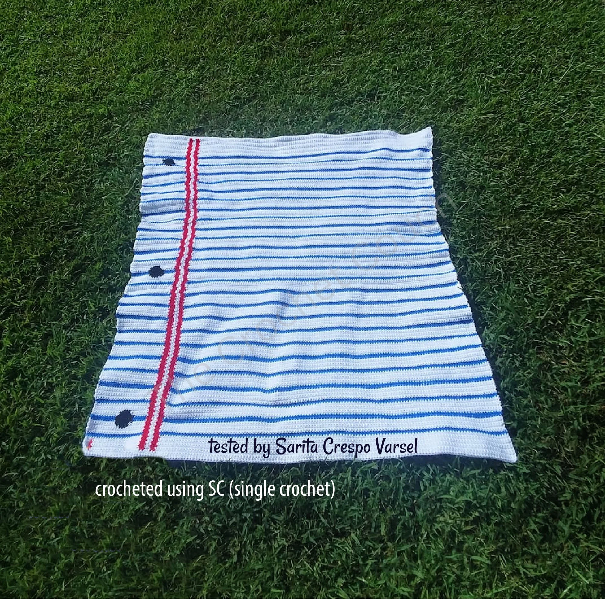A crochet afghan designed to resemble notebook paper, with white as the background, blue horizontal lines, red vertical stripes for margins, and small black "paper hole" details, laid out on grass.