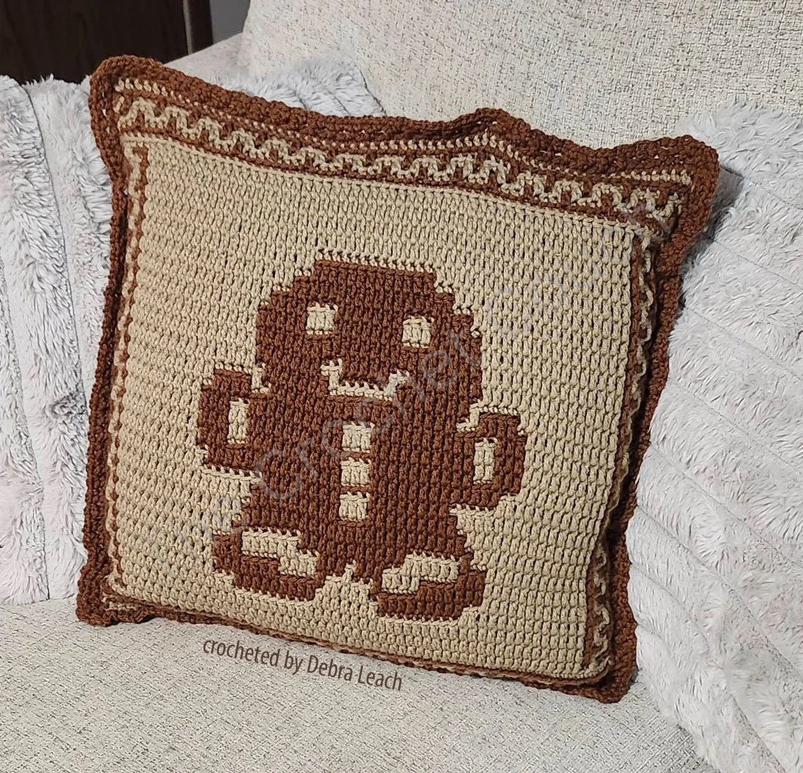 A cozy crochet pillow featuring a brown and beige Gingerbread Man design with festive details, displayed on a textured couch.