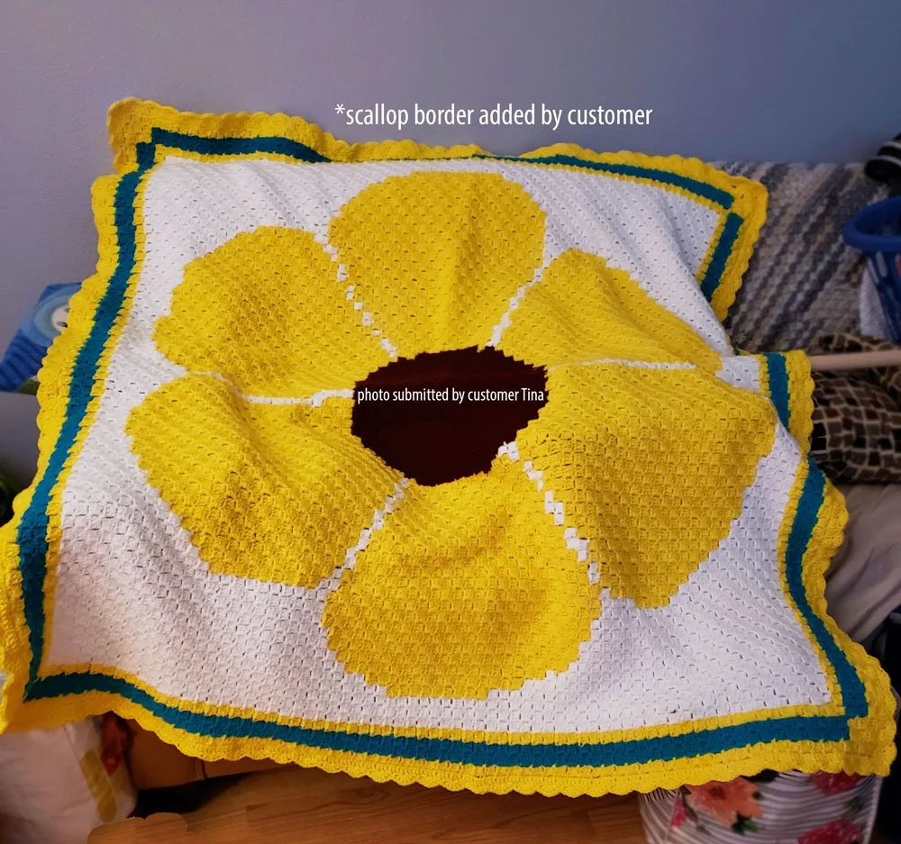 Corner to corner crochet afghan featuring a large yellow flower with a dark brown center on a white background. Edged with teal and yellow stripes and a scalloped border added by customer Tina.