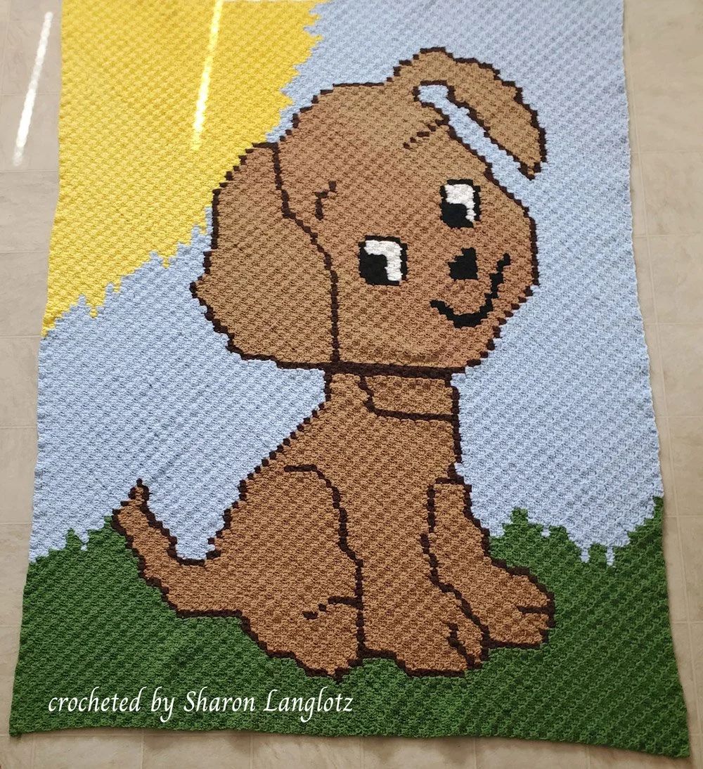 A handmade twin-bed-sized crochet afghan featuring a cheerful brown puppy sitting on green grass against a blue sky with a bright yellow sun.
