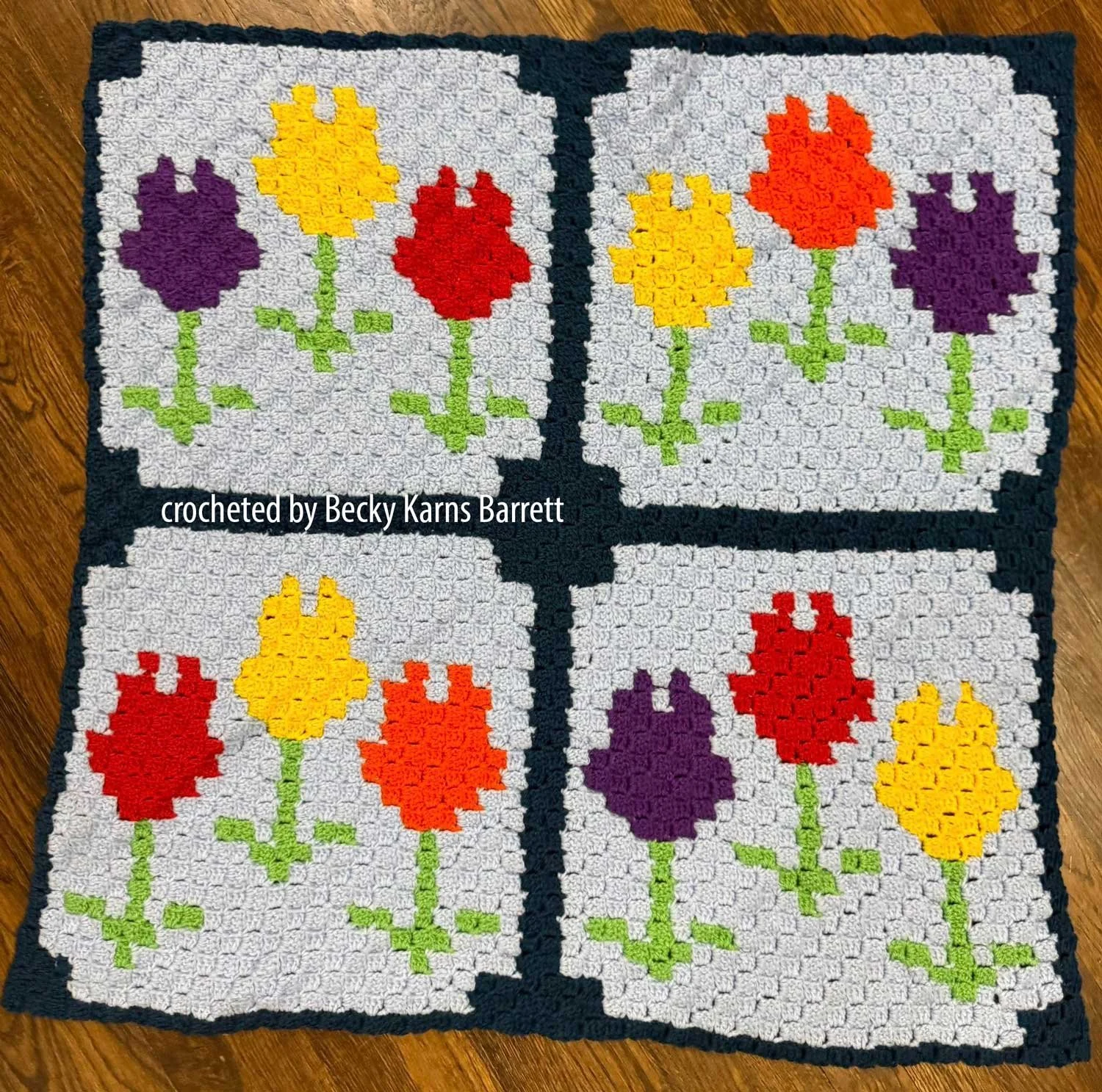 Crochet throw blanket with four blocks featuring colorful tulips in red, yellow, orange, and purple on light gray backgrounds, joined with dark borders. The layout creates a quilt-like floral design.