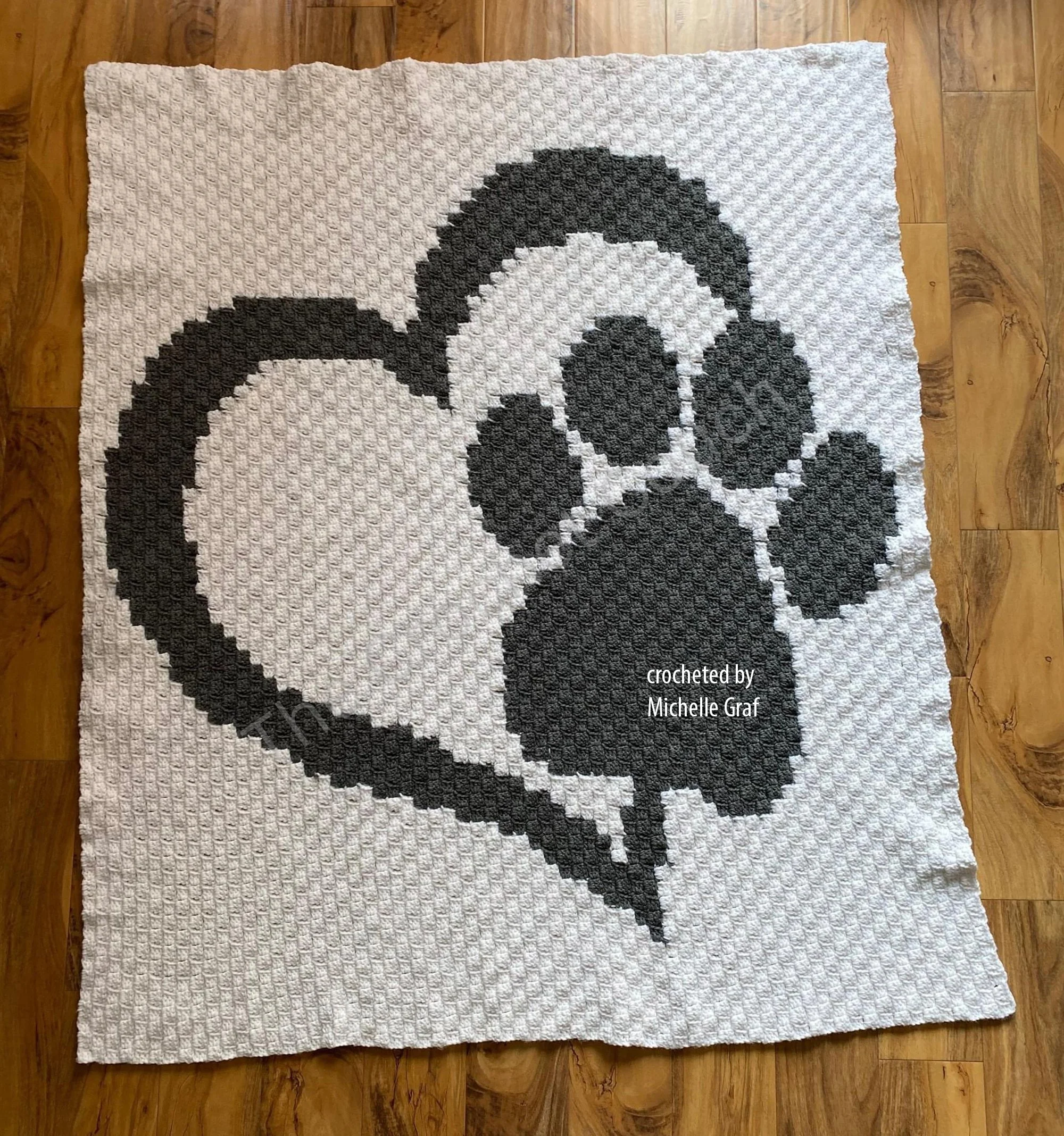 Gray and white crocheted paw in heart afghan laid flat on a wood floor, featuring a large paw print centered inside a heart shape with clean, high contrast stitches.