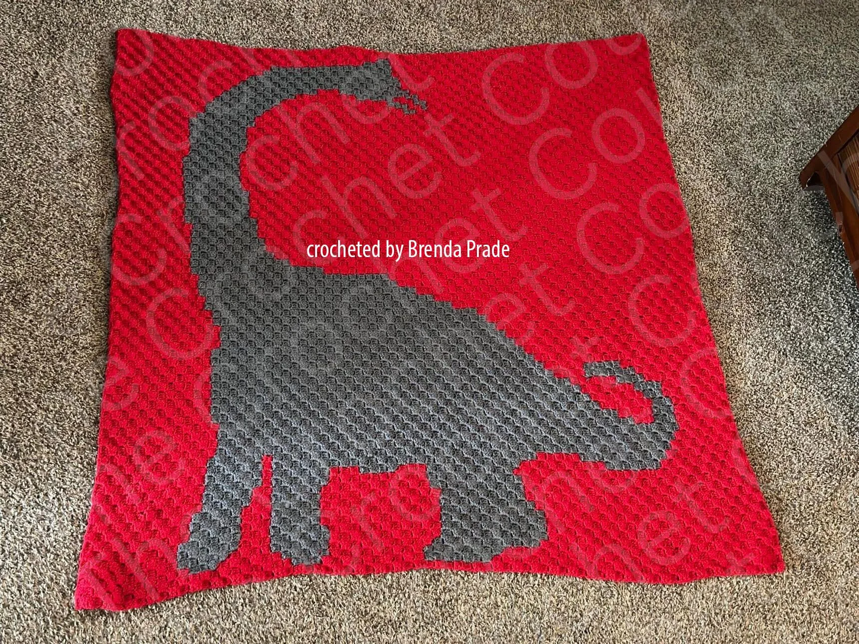 A red corner-to-corner crochet blanket featuring a large gray Brachiosaurus silhouette, laid flat on a carpeted floor.