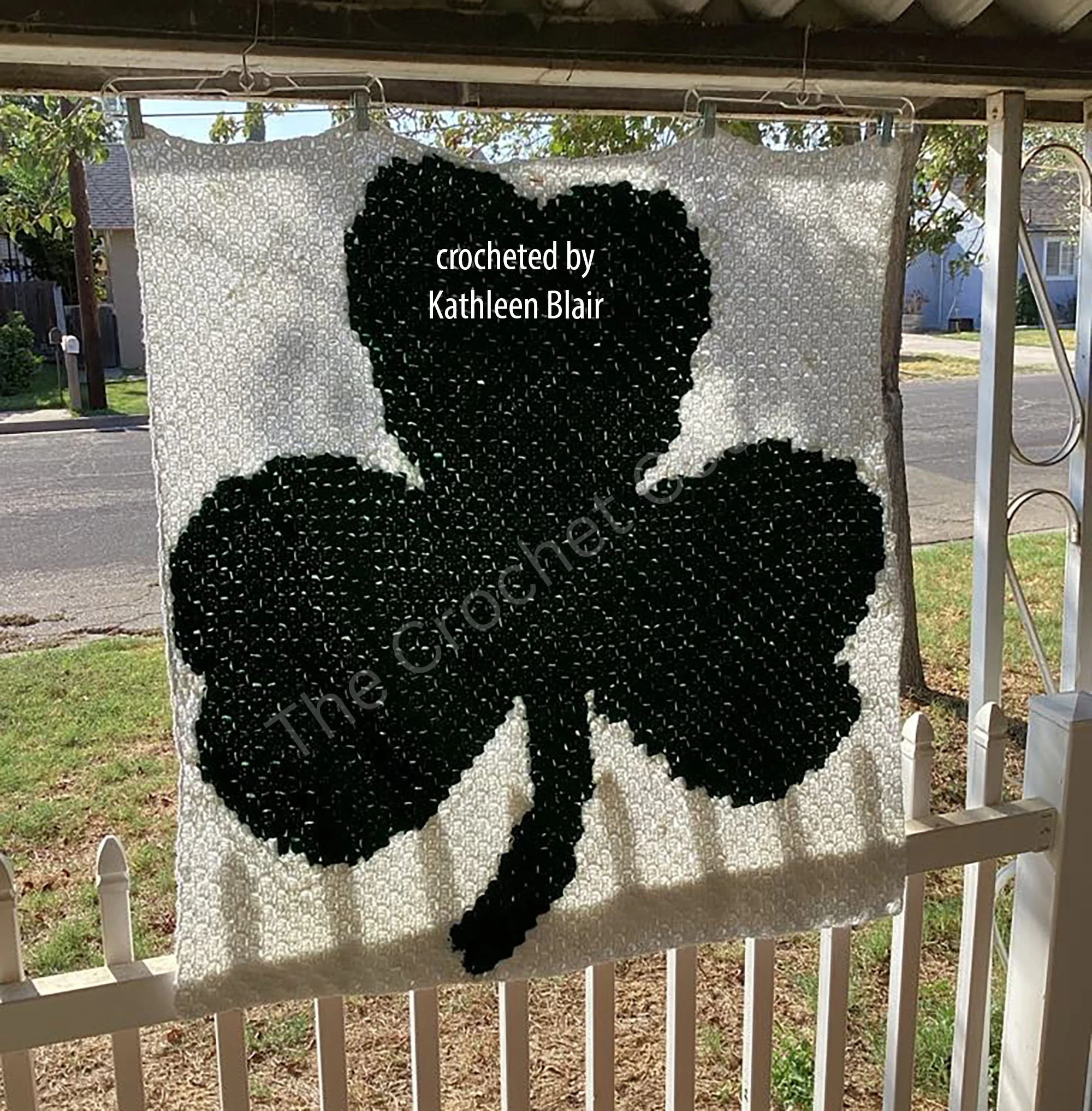 A crocheted afghan featuring a large dark green shamrock on a white background, hung outside on a porch. Crocheted by Kathleen Blair.