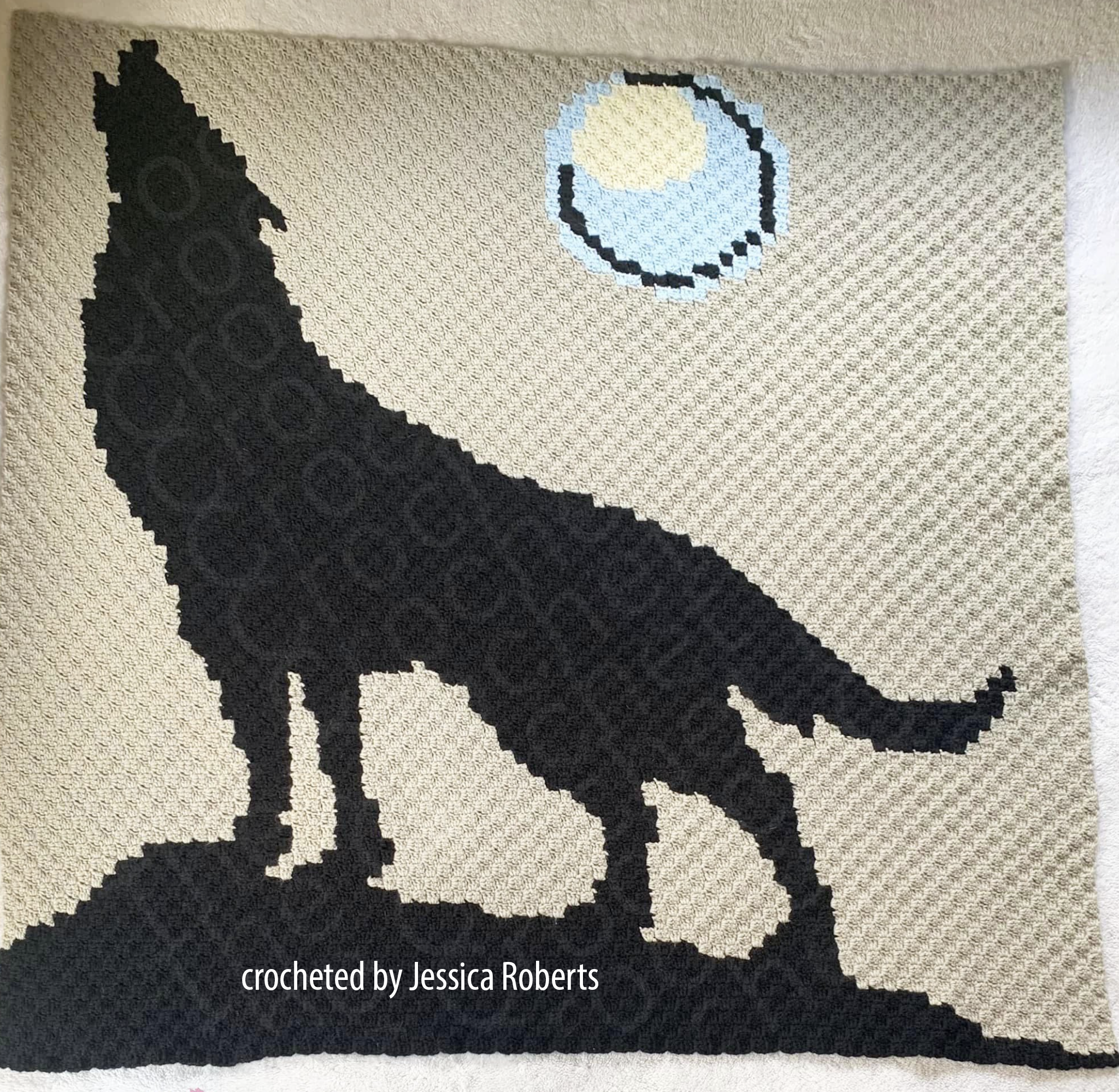 A crocheted afghan featuring a large black wolf howling at the moon against a beige background.