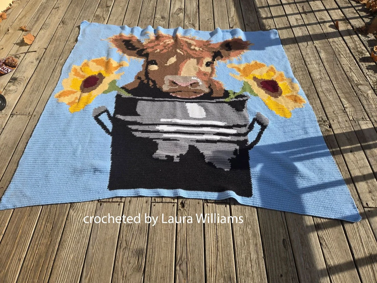 A beautifully crocheted afghan featuring a Highland cow peeking from a bucket, accented with bright sunflowers on a blue background.
