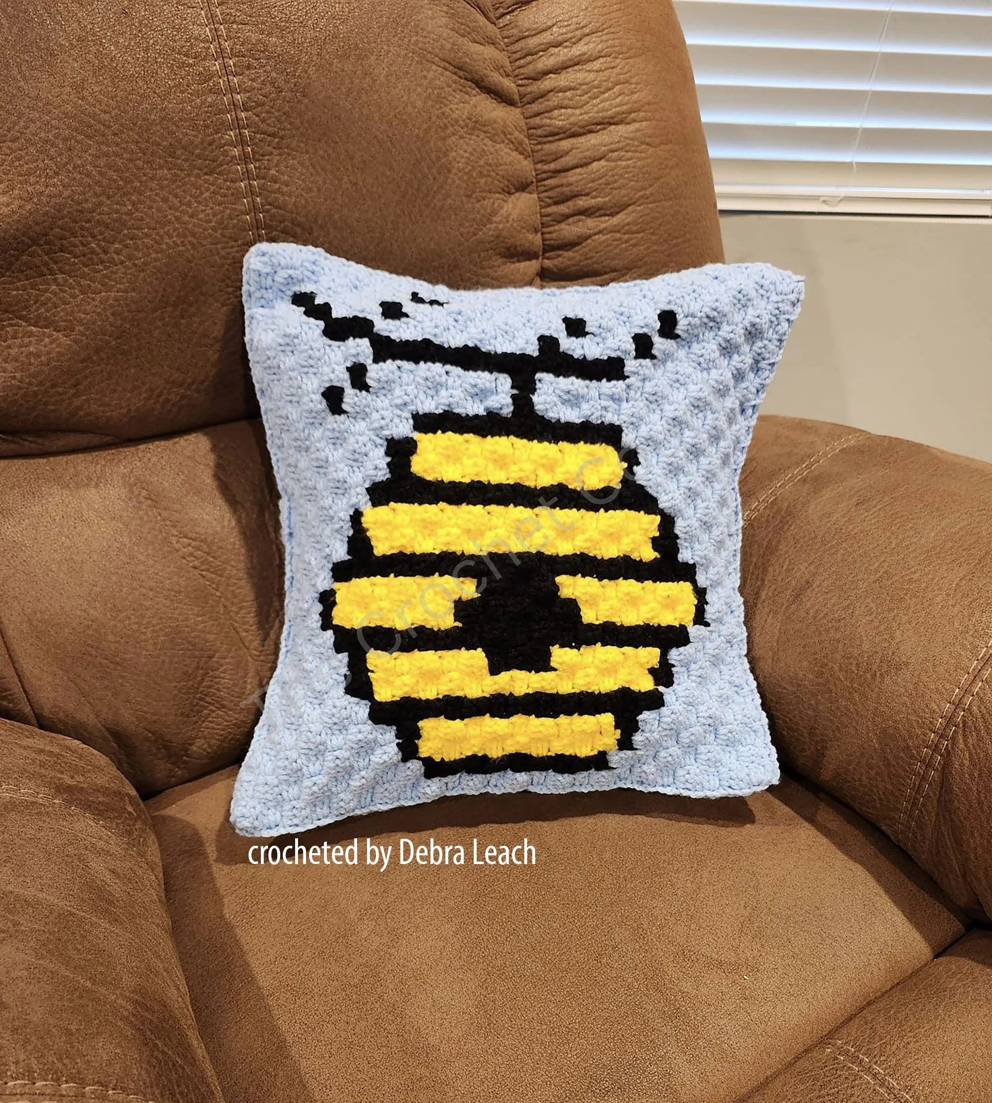 A crochet pillow featuring a beehive design in golden honey and black, set against a turquoise background.