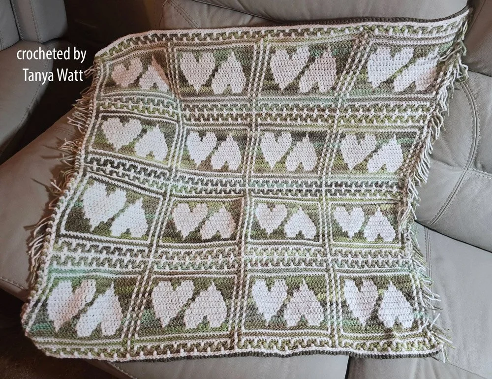 Crocheted baby afghan made of white heart motifs inside bordered squares with green and taupe backgrounds.