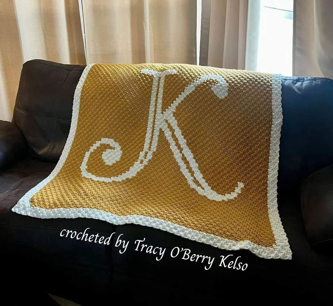 A golden mustard C2C crochet afghan featuring a large white letter K with a scalloped white border, draped over a dark leather couch.