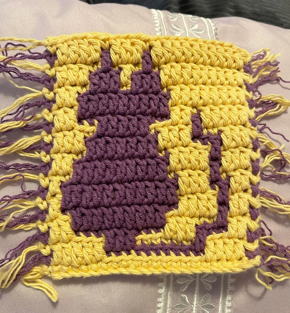 Crocheted cat mug rug in yellow and purple yarn with a pixel-style cat silhouette in the center and fringes on both sides, displayed on a lavender pillow.