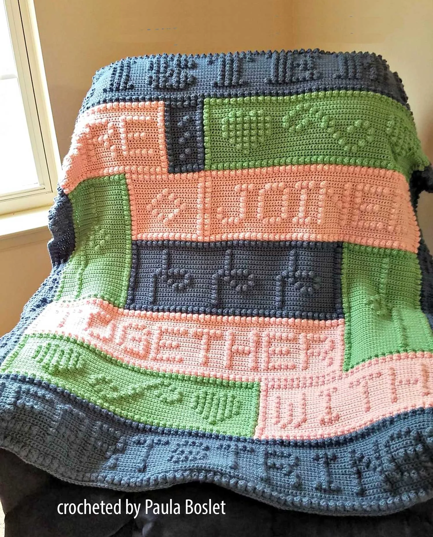 A handmade afghan worked in one piece using bobble stitch lettering and heart motifs. Panels in different colors form a rectangular layout with a wide border featuring more raised text.