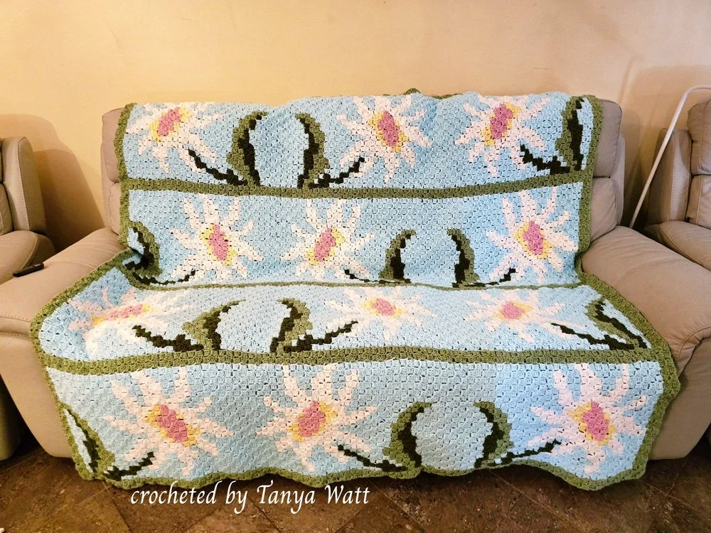 A beautifully crocheted Daisy Parade Afghan featuring delicate white daisies with pink centers and green stems on a soft blue background.