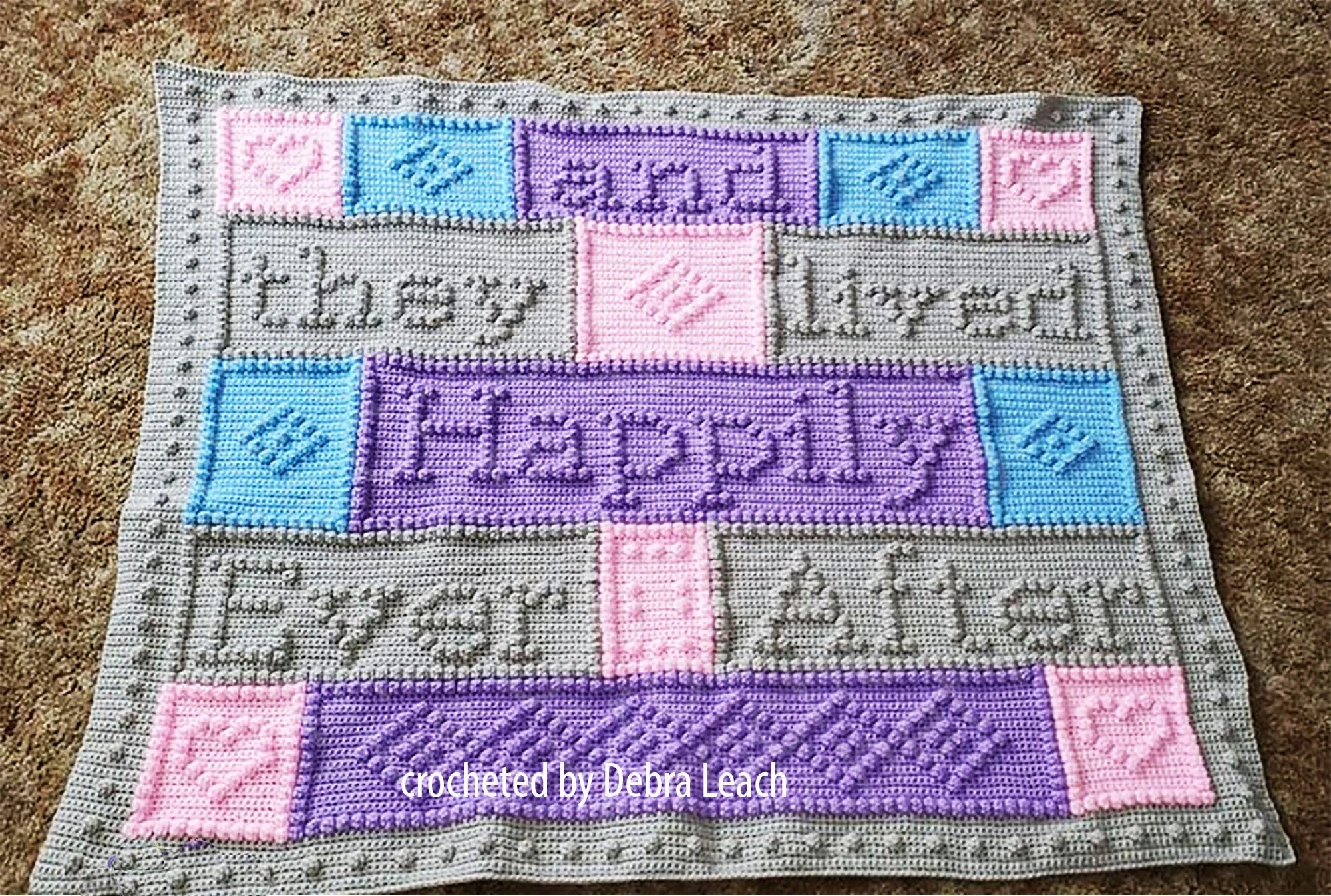 A crocheted afghan featuring the words 'And They Lived Happily Ever After' in textured bobble stitch, with pastel blocks in shades of pink, blue, purple, and grey, designed as a  wedding gift