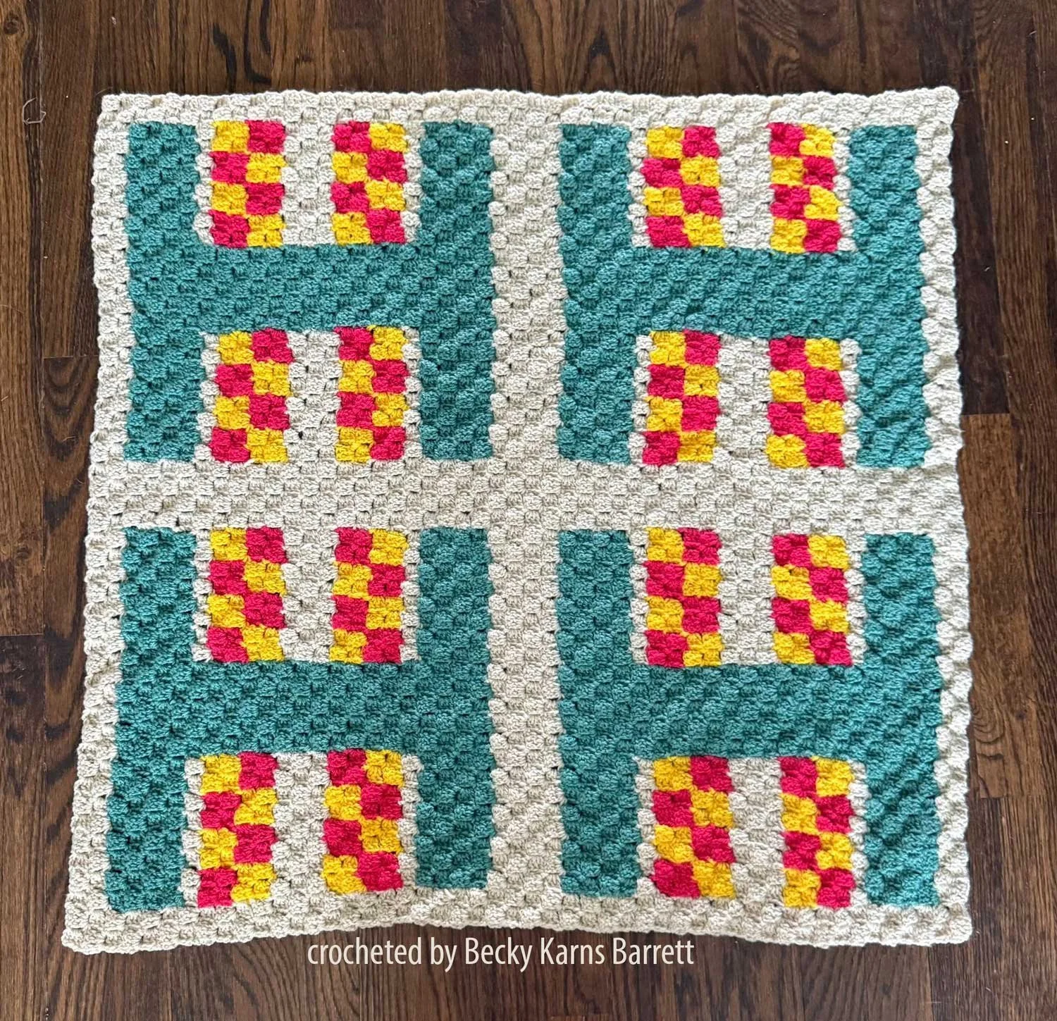 Square afghan with four repeating blocks. Deep teal “H” shapes form frames around checkerboard accents in coral and butter yellow. Background is very pale aqua for a light coastal look.