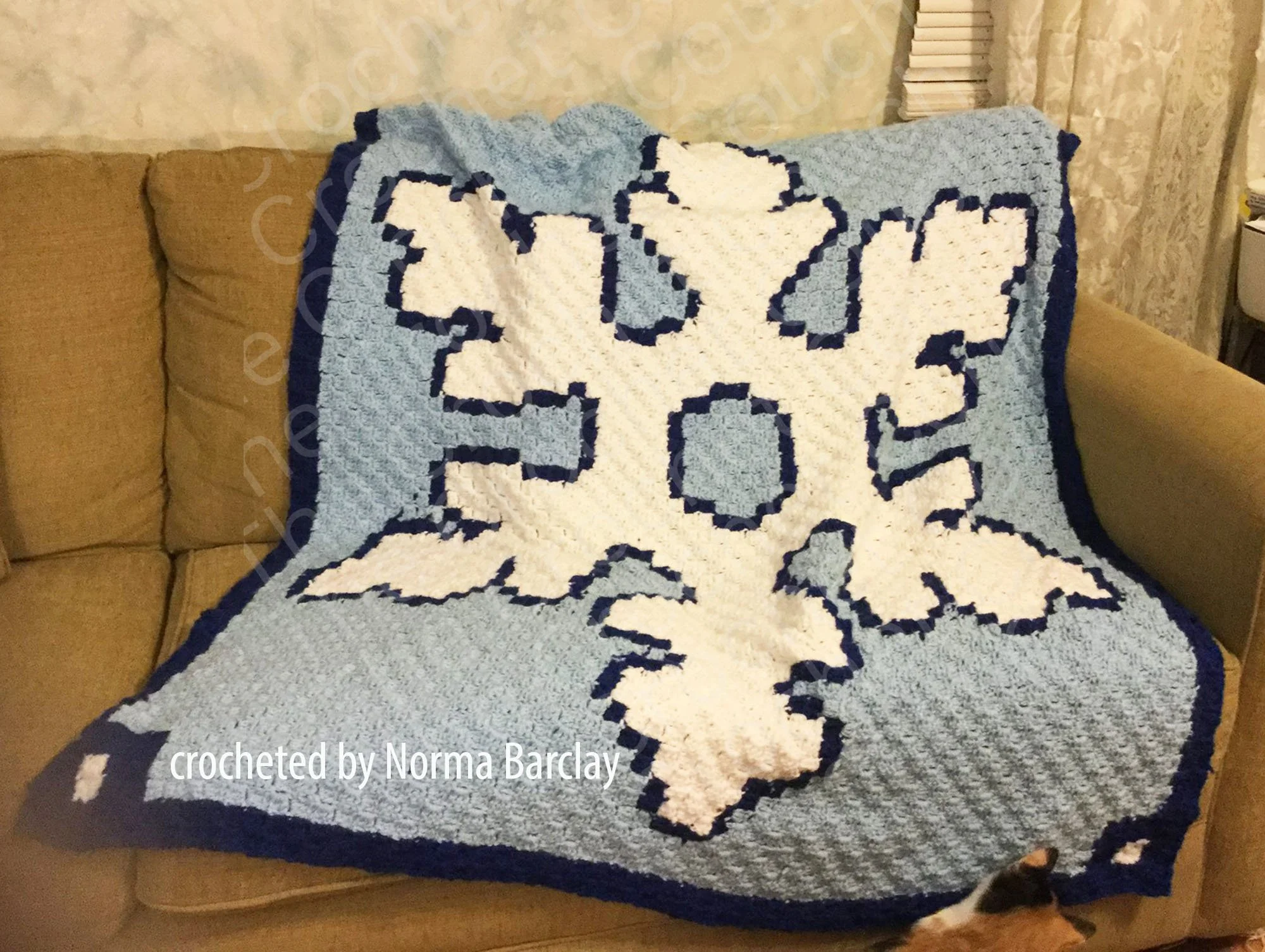 A cozy blue and white crocheted Snowflake Puffy Afghan draped on a beige sofa, featuring a large, bold snowflake design made using the C2C stitch.