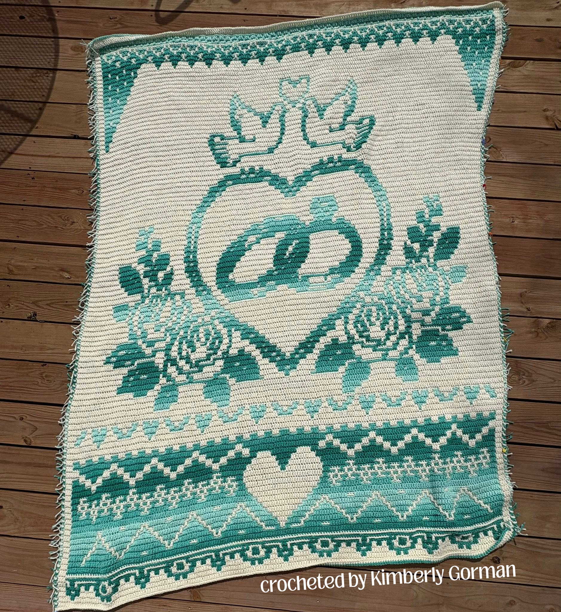 A large overlay mosaic crochet afghan in cream and teal featuring a heart with interlocking rings, floral details, and geometric borders, displayed flat on a wooden deck.