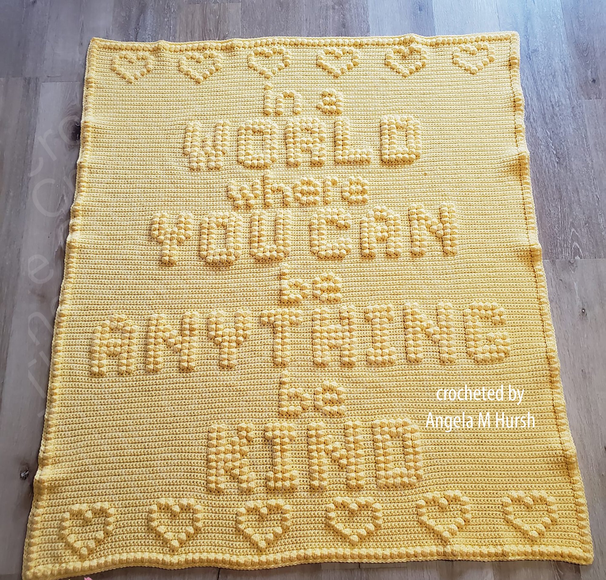 A yellow crochet afghan featuring the phrase 'In a World Where You Can Be Anything, Be Kind' with textured lettering and heart designs, laid flat on a wooden floor.