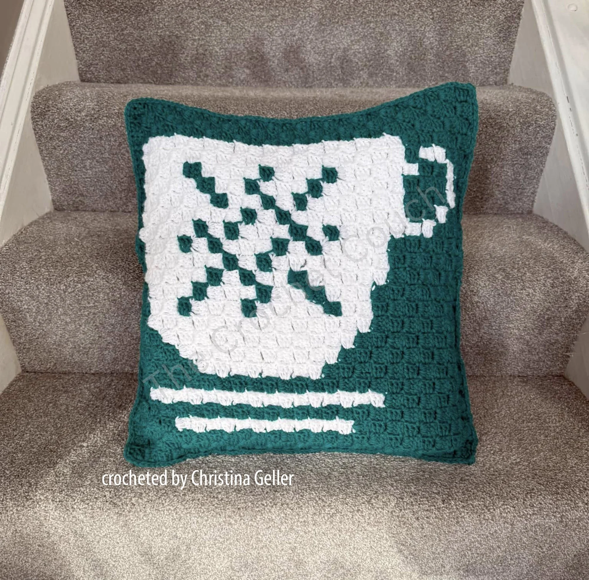 A crochet pillow featuring a white teacup design on a green background, and is displayed on a staircase, showcasing its cozy charm as part of a three-design teacup pillow collection.