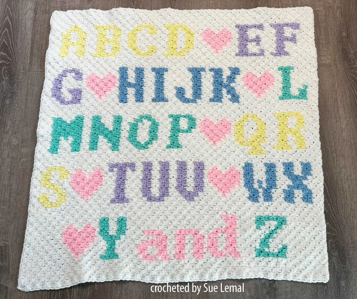 Baby afghan laying on floor with pastel colored letters on a white background