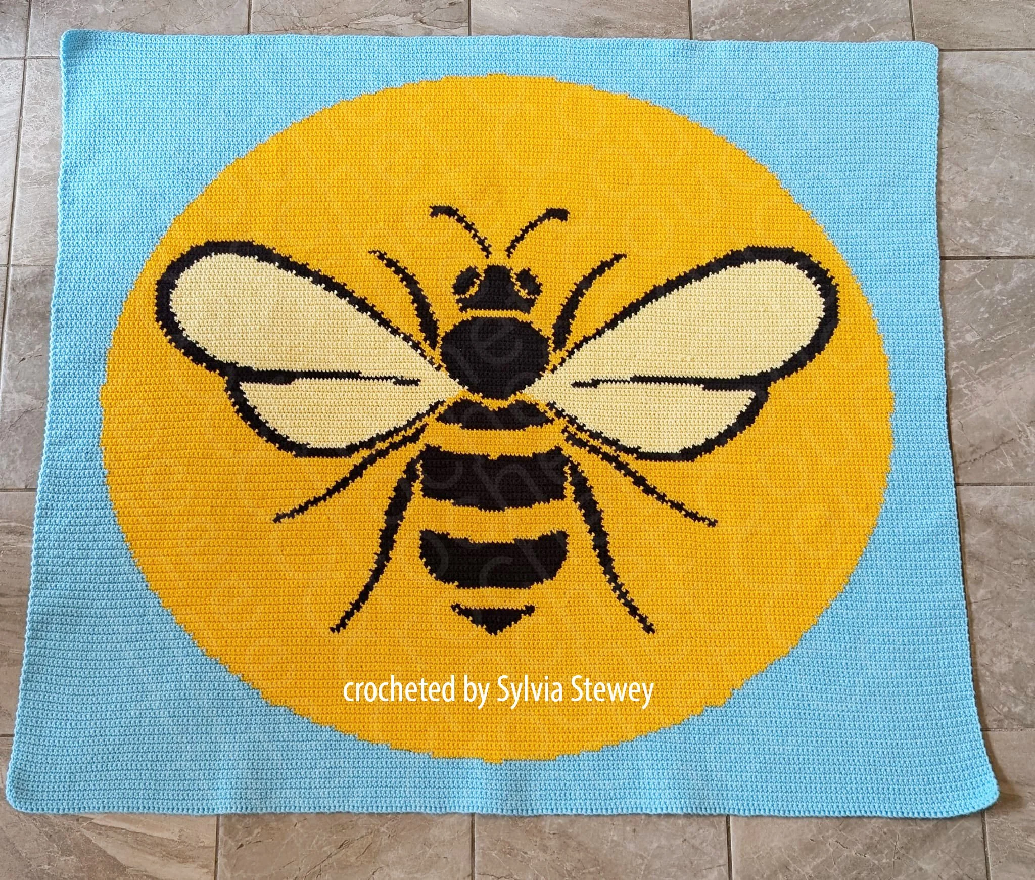 A single crochet afghan featuring a large, detailed bee in front of a yellow circle, set against a blue background, laid flat on a tiled floor.