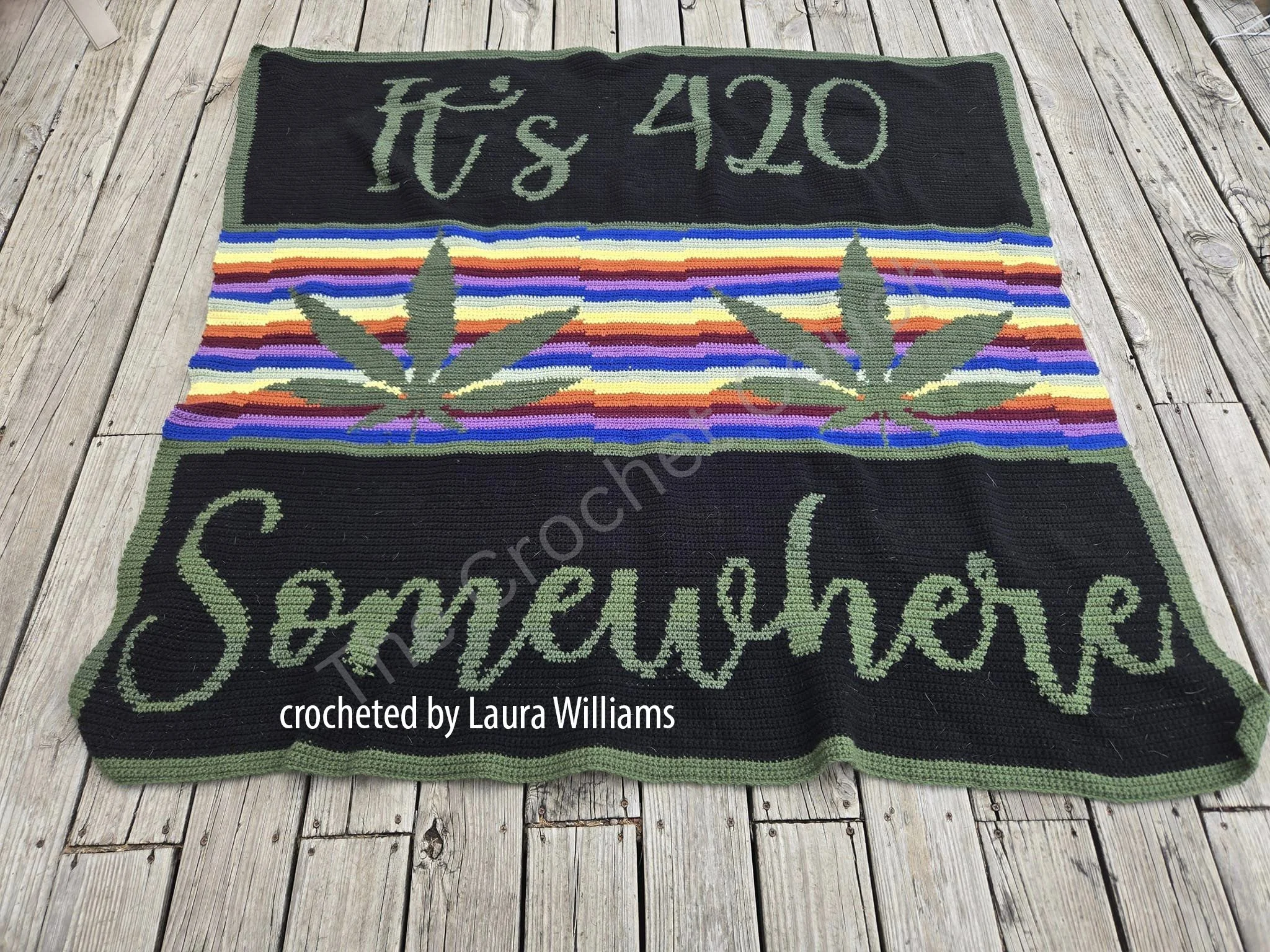 Crocheted afghan with the words "It's 420 Somewhere" in green, featuring cannabis leaves and colorful rainbow stripes, displayed on a wooden deck.