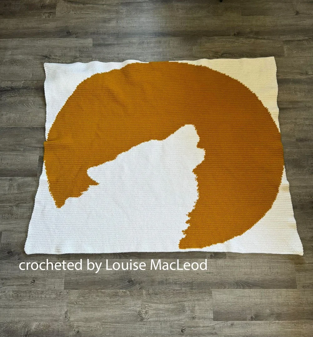 A handmade crochet afghan featuring a bold silhouette of a howling wolf against a large full moon, crafted in warm tones.