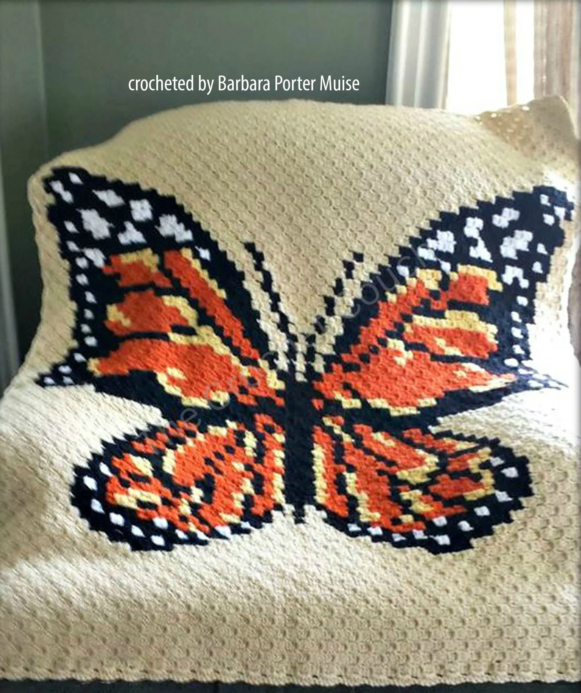 A stunning crochet afghan featuring a large, vibrant Monarch butterfly with intricate wing details. The afghan is displayed draped over a chair.