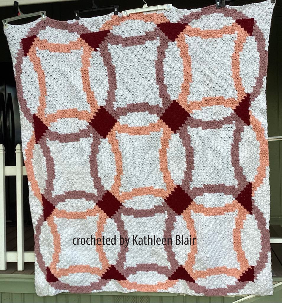 Corner to Corner crochet afghan with an interlocking wedding ring design in white, peach, mauve, and burgundy tones, hanging outdoors against a porch railing.