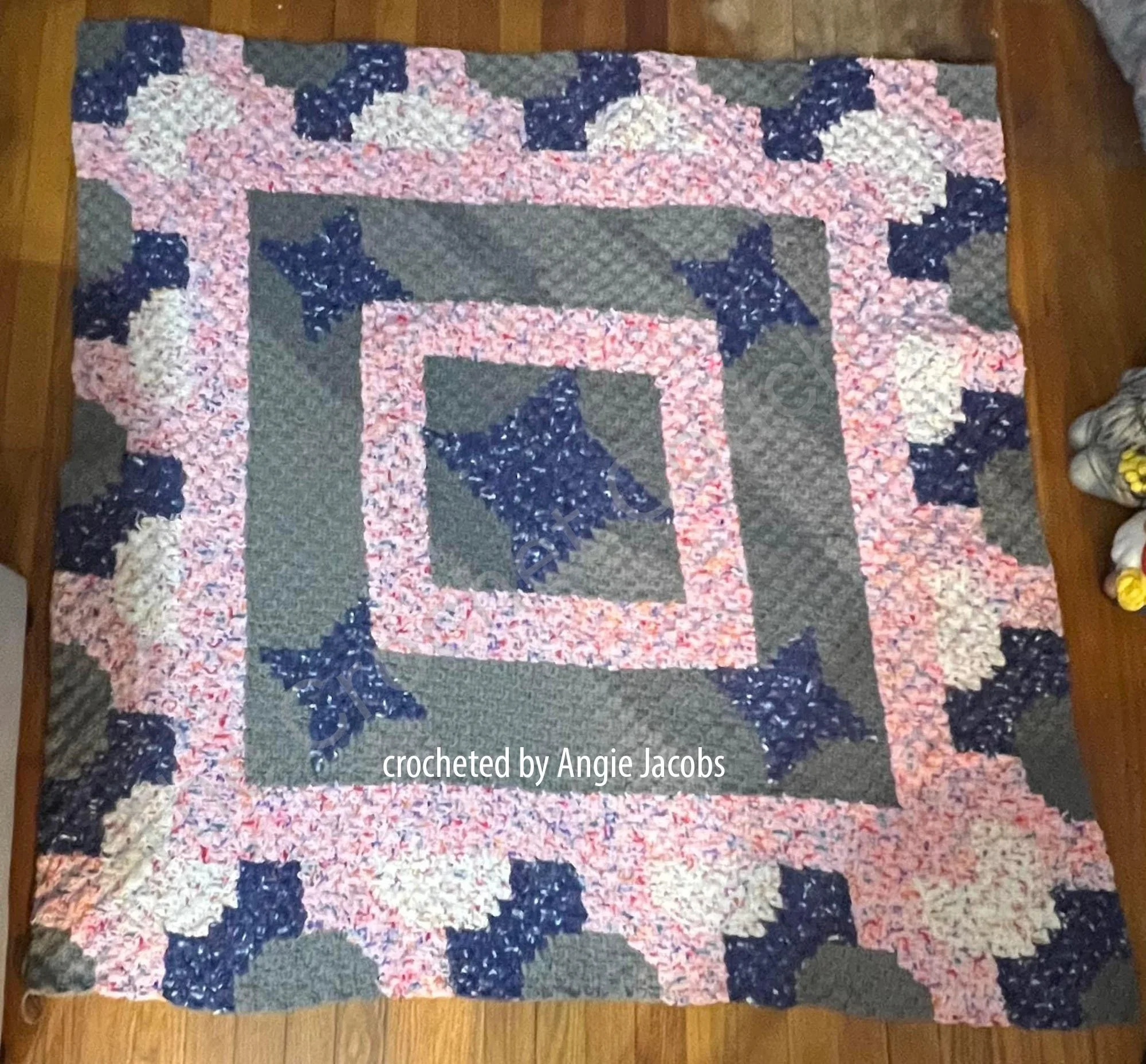 A completed Bowtie Bounce Afghan crocheted in shades of blue, pink, gray, and white, with a bold geometric bowtie design. The afghan is displayed on a wooden floor, crocheted by Angie Jacobs.
