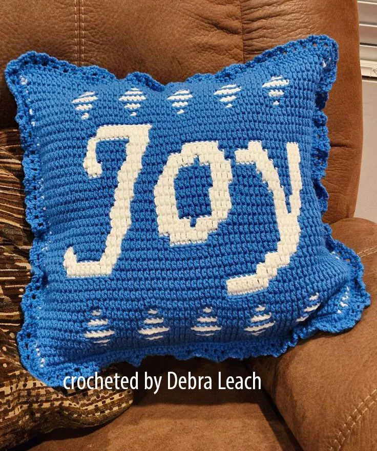 A vibrant blue crochet pillow with the word "Joy" stitched in white, featuring charming details and a lacy edge, displayed on a cozy brown chair.