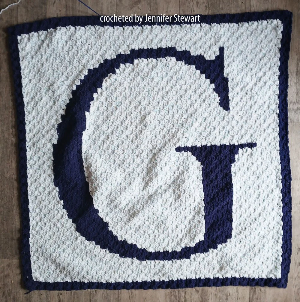Corner to corner crochet afghan featuring a large, dark blue letter "G" on a light background with matching dark border, laid flat on wooden floor. Crocheted by Jennifer Stewart.