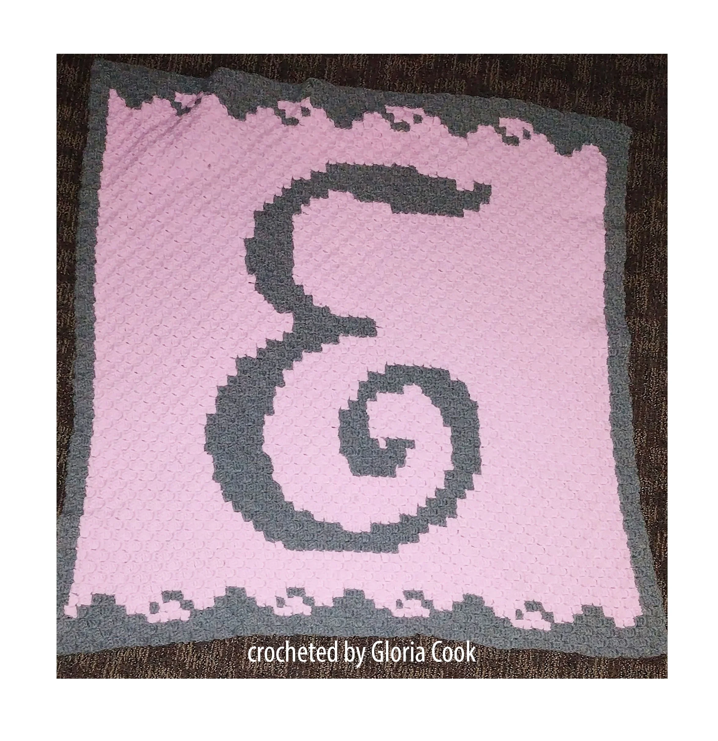 A crocheted baby afghan featuring the letter "E" in gray against a pink background, bordered with a matching gray trim.