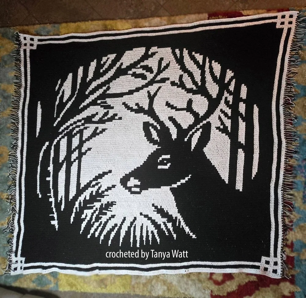 Crocheted afghan featuring a bold black and white silhouette of a deer surrounded by trees, with a geometric border. Crocheted by Tanya Watt.
