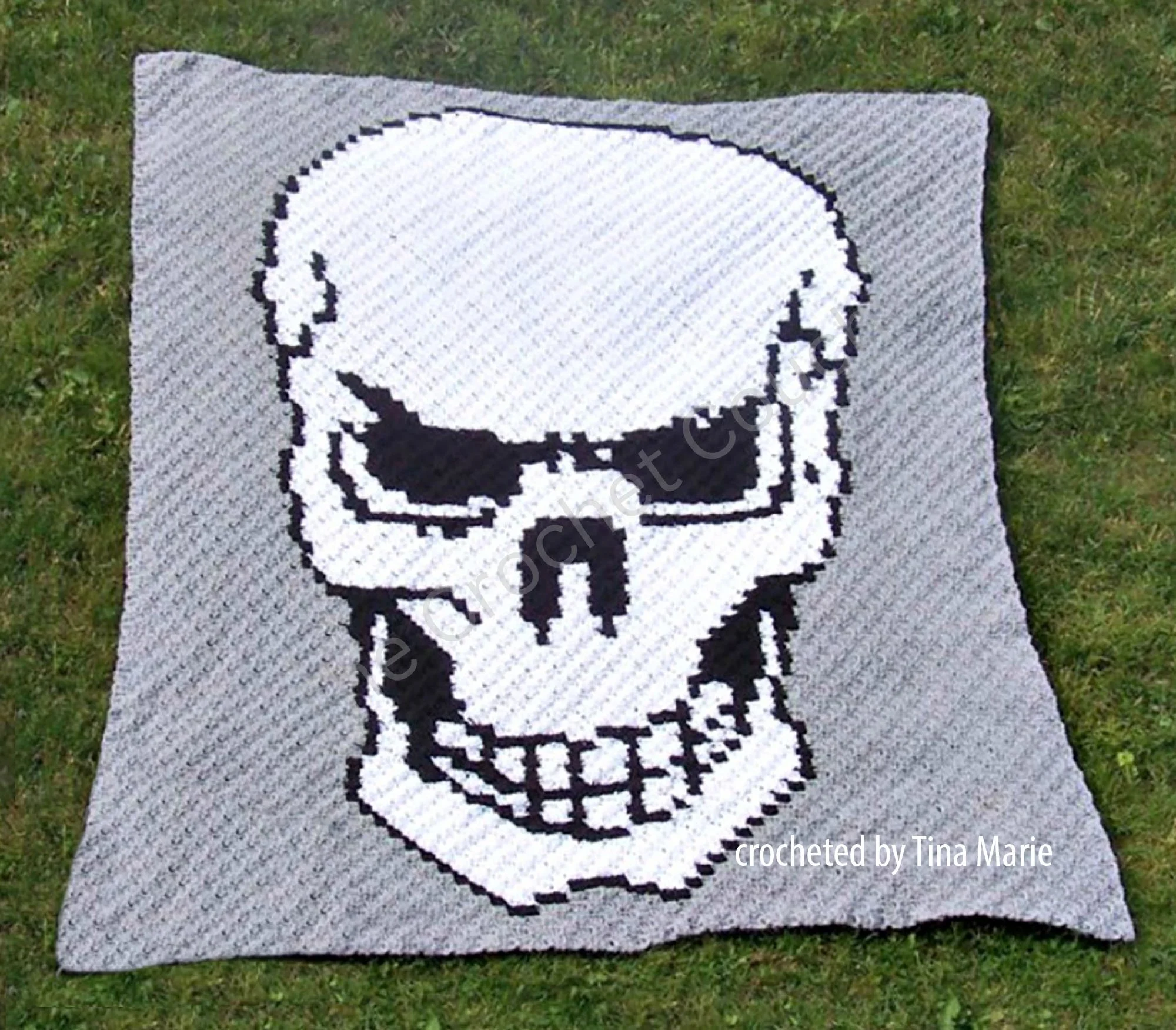 A crocheted afghan with a large white skull design outlined in black, set against a grey background, made by Tina Marie. The afghan is displayed on green grass.