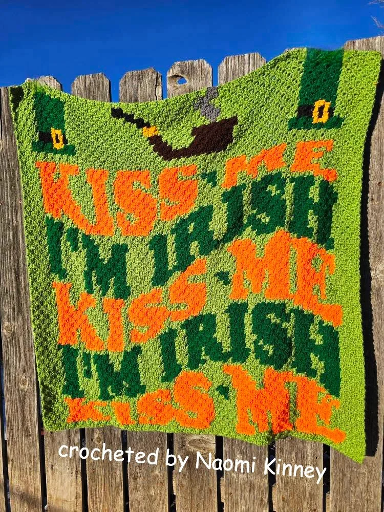 A St. Patrick’s Day crochet afghan featuring the words “Kiss Me, I’m Irish” in bold orange and green letters, along with festive leprechaun hats and a pot of gold. Crocheted by Naomi Kinney.