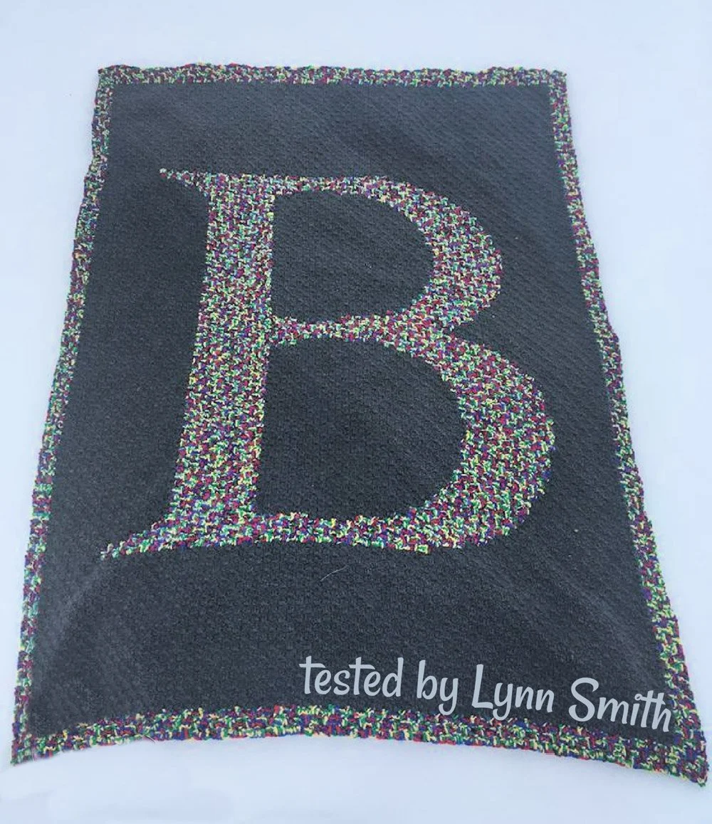 Crochet afghan featuring a large uppercase letter B centered on a dark gray background, with a colorful multi-tone yarn outlining the letter and forming the border.